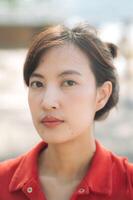 Asian thoughtful woman in red shirt gazes directly at camera, with soft natural light highlighting her features photo