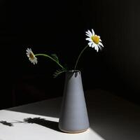 A single white daisy in modern dark gray ceramic vase illuminated by moody lighting, creating an elegant and artistic still life composition photo