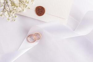 Elegant wedding background in a white tone. Two gold wedding rings lie on the spiral of a satin ribbon. flowers on a wedding envelope. top view photo