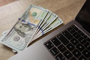A stack of hundred dollar bills placed on a wooden surface next to a laptop keyboard. Concept of online earnings, digital finance and remote work. photo