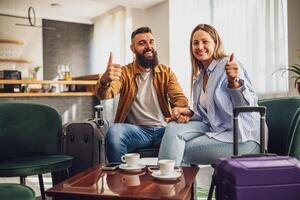 Married couple arrived in hotel. They are on vacation, sitting in lobby with suitcases, looking at camera, smiling and showing thumbs up. photo