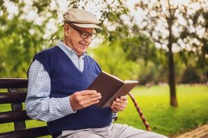 Old man with hat is sitting on bench in park on sunny day. He is relaxing and using reading a book. photo