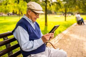 Old man with hat is sitting on bench in park on sunny day and using digital tablet. photo