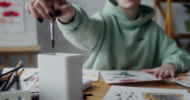 A young female artist washes a drawing brush in water and continues to paint a flower with watercolor paint on paper, while working in a workshop video