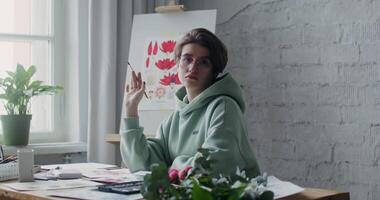 Young female artist in glasses looking at the camera, while posing with a drawing brush in her hands while sitting at a desk with drawings laid out on it video