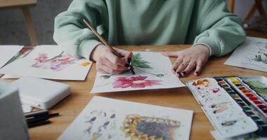 Close-up of female hands painting with watercolors in the workshop. Sheets of paper with painted flowers spread out on the table video