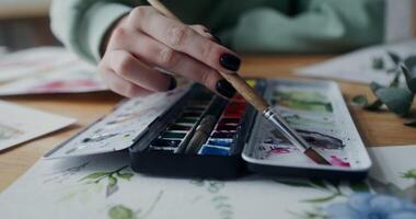 A woman artist mixes watercolor paints with a brush to get the right color. Close-up of female hands with a paintbrush while working in a workshop video