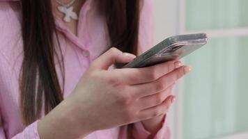 Woman holding a smartphone with both hands while wearing a pink shirt, close-up view of fingers interacting with the device in a calm indoor setting video