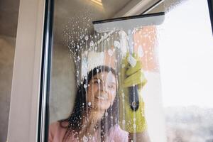 Smiling housekeeper cleaning window with squeegee and detergent photo