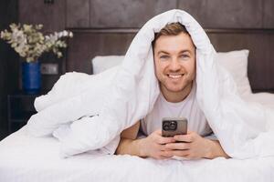 Man lying in bed under blanket using smartphone photo