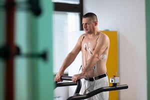 A middle-aged man is undergoing a medical stress test on a treadmill, connected to ECG electrodes for heart monitoring in a clinical environment photo