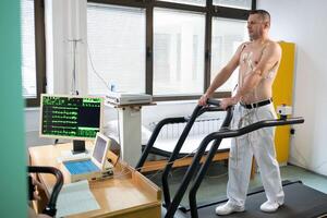 A middle-aged man is undergoing a medical stress test on a treadmill, connected to ECG electrodes for heart monitoring in a clinical environment photo