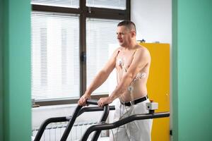 A middle-aged man is undergoing a medical stress test on a treadmill, connected to ECG electrodes for heart monitoring in a clinical environment photo