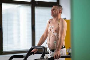 A middle-aged man is undergoing a medical stress test on a treadmill, connected to ECG electrodes for heart monitoring in a clinical environment photo