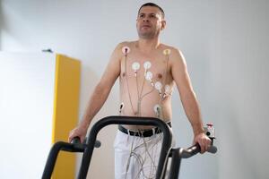 A middle-aged man is undergoing a medical stress test on a treadmill, connected to ECG electrodes for heart monitoring in a clinical environment photo