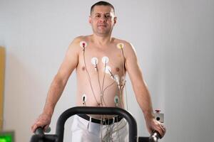 A middle-aged man is undergoing a medical stress test on a treadmill, connected to ECG electrodes for heart monitoring in a clinical environment photo