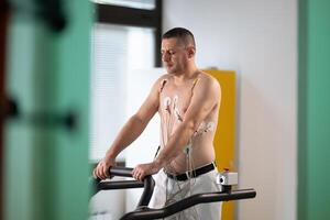 A middle-aged man is undergoing a medical stress test on a treadmill, connected to ECG electrodes for heart monitoring in a clinical environment photo