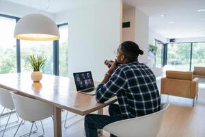 African American businessman in glasses sitting at a table in a modern living room, using a laptop and smartphone for business chat, conversation with friends and entertainment. photo