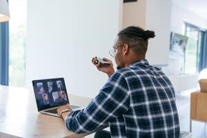 African American businessman in glasses sitting at a table in a modern living room, using a laptop and smartphone for business chat, conversation with friends and entertainment. photo