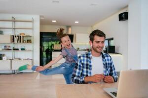 Work from home. Daughter interrupting her father while he is having a business online conversation on his laptop while sitting in the modern living room. photo
