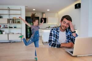 Work from home. Daughter interrupting her father while he is having a business online conversation on his laptop while sitting in the modern living room. photo