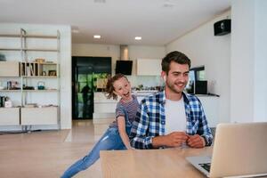 Work from home. Daughter interrupting her father while he is having a business online conversation on his laptop while sitting in the modern living room. photo