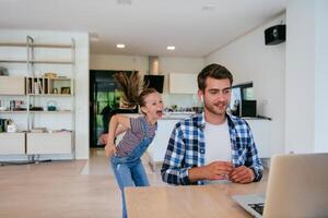 Work from home. Daughter interrupting her father while he is having a business online conversation on his laptop while sitting in the modern living room. photo