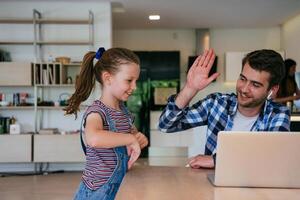 Work from home. Daughter interrupting her father while he is having a business online conversation on his laptop while sitting in the modern living room. photo