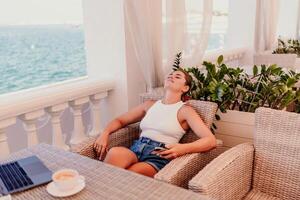 woman is sitting at a table with a laptop and a cup of coffee. She is wearing sunglasses and a ponytail. The scene suggests a relaxed and leisurely atmosphere photo