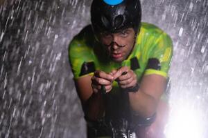 A triathlete braving the rain as he cycles through the night, preparing himself for the upcoming marathon. The blurred raindrops in the foreground and the dark, moody atmosphere in the background add photo