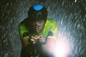 A triathlete braving the rain as he cycles through the night, preparing himself for the upcoming marathon. The blurred raindrops in the foreground and the dark, moody atmosphere in the background add photo