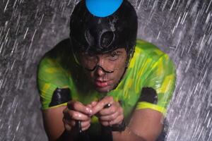 A triathlete braving the rain as he cycles through the night, preparing himself for the upcoming marathon. The blurred raindrops in the foreground and the dark, moody atmosphere in the background add photo