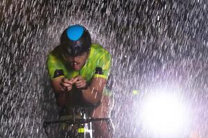 A triathlete braving the rain as he cycles through the night, preparing himself for the upcoming marathon. The blurred raindrops in the foreground and the dark, moody atmosphere in the background add photo