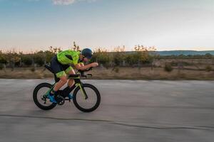 Triathlete riding his bicycle during sunset, preparing for a marathon. The warm colors of the sky provide a beautiful backdrop for his determined and focused effort. photo