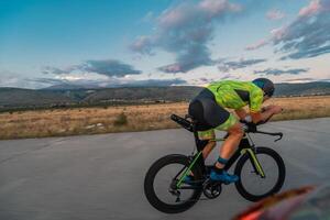 Triathlete riding his bicycle during sunset, preparing for a marathon. The warm colors of the sky provide a beautiful backdrop for his determined and focused effort. photo