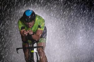 A triathlete braving the rain as he cycles through the night, preparing himself for the upcoming marathon. The blurred raindrops in the foreground and the dark, moody atmosphere in the background add photo