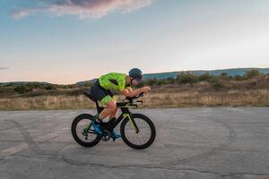 Triathlete riding his bicycle during sunset, preparing for a marathon. The warm colors of the sky provide a beautiful backdrop for his determined and focused effort. photo