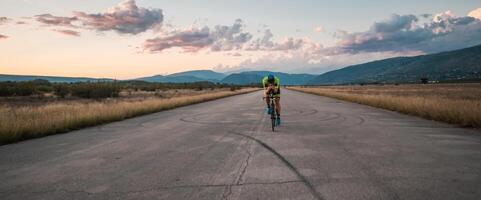 Triathlete riding his bicycle during sunset, preparing for a marathon. The warm colors of the sky provide a beautiful backdrop for his determined and focused effort. photo
