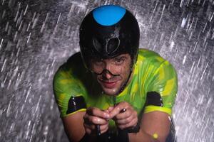 A triathlete braving the rain as he cycles through the night, preparing himself for the upcoming marathon. The blurred raindrops in the foreground and the dark, moody atmosphere in the background add photo