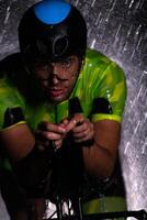 A triathlete braving the rain as he cycles through the night, preparing himself for the upcoming marathon. The blurred raindrops in the foreground and the dark, moody atmosphere in the background add photo
