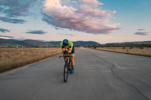 Triathlete riding his bicycle during sunset, preparing for a marathon. The warm colors of the sky provide a beautiful backdrop for his determined and focused effort. photo