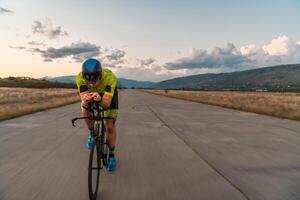 Triathlete riding his bicycle during sunset, preparing for a marathon. The warm colors of the sky provide a beautiful backdrop for his determined and focused effort. photo