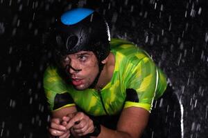 A triathlete braving the rain as he cycles through the night, preparing himself for the upcoming marathon. The blurred raindrops in the foreground and the dark, moody atmosphere in the background add photo