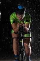 A triathlete braving the rain as he cycles through the night, preparing himself for the upcoming marathon. The blurred raindrops in the foreground and the dark, moody atmosphere in the background add photo