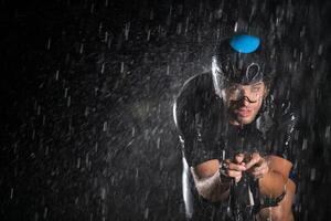 A triathlete braving the rain as he cycles through the night, preparing himself for the upcoming marathon. The blurred raindrops in the foreground and the dark, moody atmosphere in the background add photo