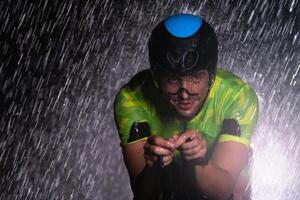 A triathlete braving the rain as he cycles through the night, preparing himself for the upcoming marathon. The blurred raindrops in the foreground and the dark, moody atmosphere in the background add photo