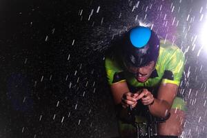 A triathlete braving the rain as he cycles through the night, preparing himself for the upcoming marathon. The blurred raindrops in the foreground and the dark, moody atmosphere in the background add photo