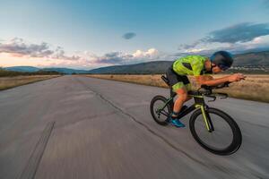 Triathlete riding his bicycle during sunset, preparing for a marathon. The warm colors of the sky provide a beautiful backdrop for his determined and focused effort. photo