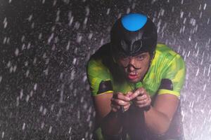 A triathlete braving the rain as he cycles through the night, preparing himself for the upcoming marathon. The blurred raindrops in the foreground and the dark, moody atmosphere in the background add photo