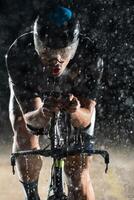 A triathlete braving the rain as he cycles through the night, preparing himself for the upcoming marathon. The blurred raindrops in the foreground and the dark, moody atmosphere in the background add photo